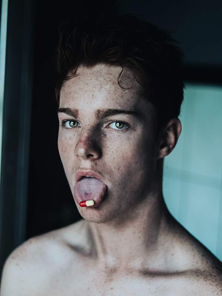 Gezicht van jonge man met capsule op uitstekende tong, met een blik van "help mij". Afhankelijkheid van medicatie ontwikkelt zich geleidelijk.