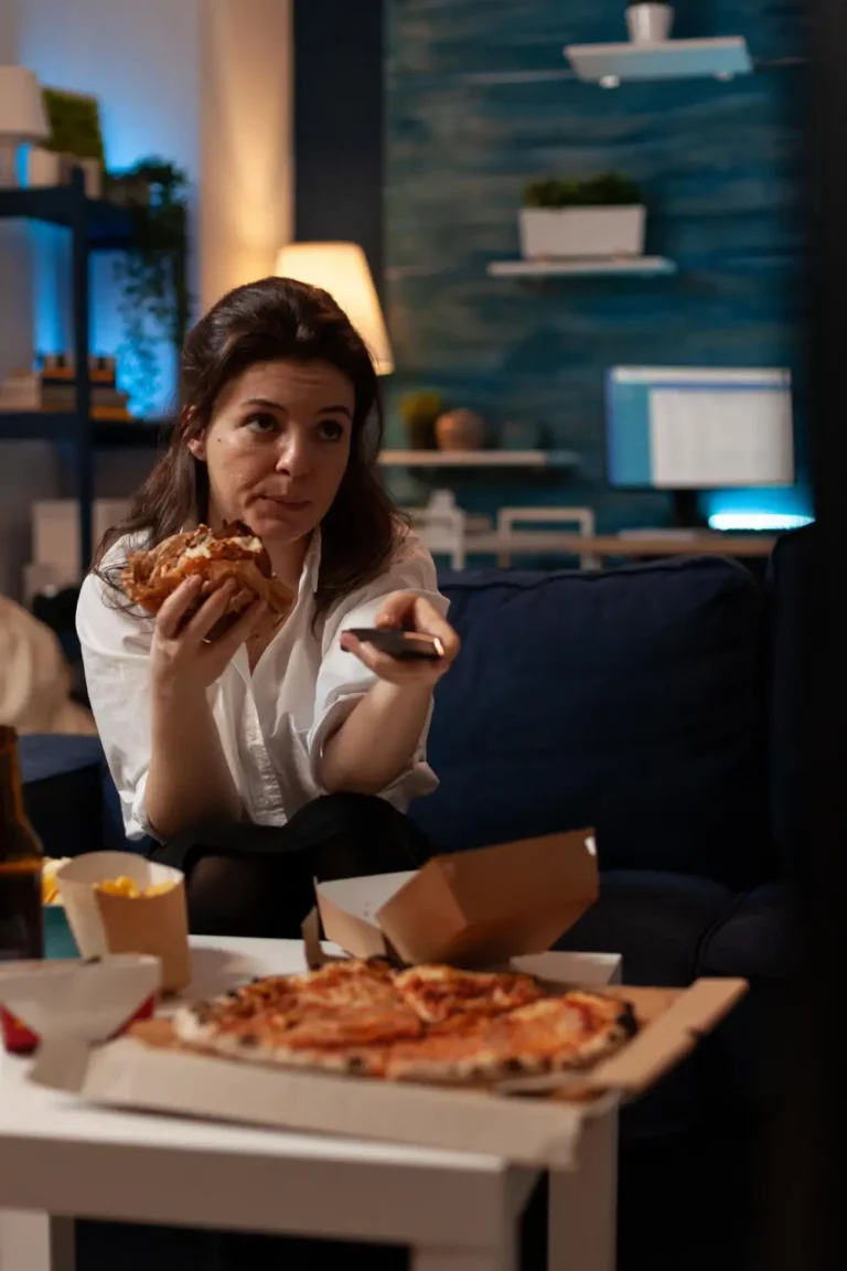 Vrouw kampt met emotioneel eten terwijl ze met hamburger en afstandsbediening in haar hand televisie kijkt.